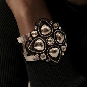 Pu&ograve; includere: Un bracciale a fascia larga con una banda in metallo color argento e una grande placca centrale decorata. La placca &egrave; composta da cornici nere che circondano forme lisce e riflettenti color argento a goccia e quadrate, con piccoli cristalli trasparenti.