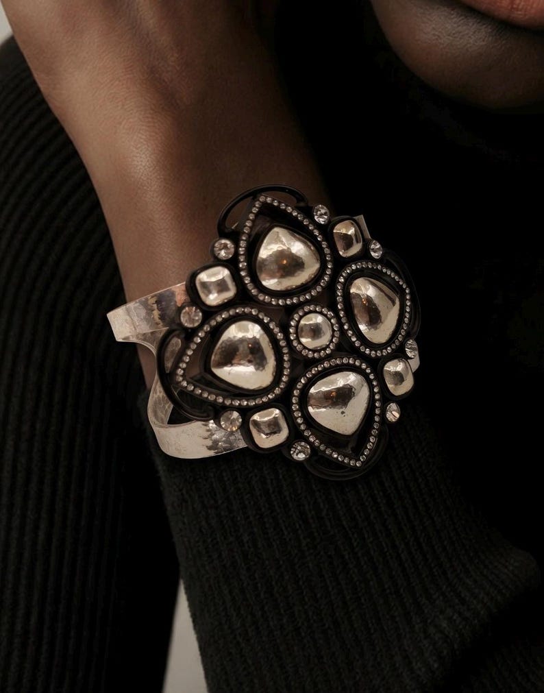 Pu&ograve; includere: Un bracciale a fascia larga con una banda in metallo color argento e una grande placca centrale decorata. La placca &egrave; composta da cornici nere che circondano forme lisce e riflettenti color argento a goccia e quadrate, con piccoli cristalli trasparenti.