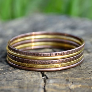 Op de afbeelding: Set van vijf gouden en koperen armbanden, gestapeld op een houten oppervlak. De armbanden hebben verschillende texturen, waaronder gehamerd, geribbeld en glad.