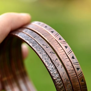 May include: Three copper bracelets with different designs. One bracelet has a hammered texture, another has a geometric pattern, and the third has a floral design.