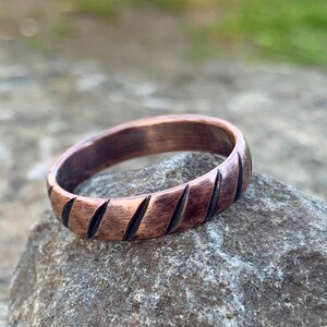 Pode incluir: Um anel de cobre com ranhuras diagonais. O anel é de cor castanha quente e está sobre uma pedra cinzenta. As ranhuras são escuras, criando um contraste com o cobre. O anel é uma joia.