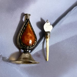 May include: A metallic perfume bottle with a teardrop-shaped amber-colored inlay and a textured silver-tone border. The bottle features a wide, flared base and a narrow neck. Beside it lies a matching silver-tone stopper with a long, thin, cream-colored applicator.