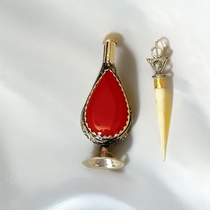 May include: A teardrop-shaped perfume bottle featuring a vibrant red center panel framed by intricate silver-toned metalwork, a gold-toned neck, and a pedestal base, accompanied by a slender, pointed applicator tool with a decorative metal top.