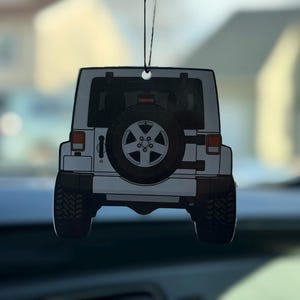 May include: White Jeep air freshener hanging from a black string. The air freshener is a detailed illustration of the rear of a white Jeep SUV, including the spare tire and black tires. The background is blurred.