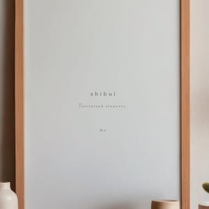 May include: A framed print with the word "shibui" and the phrase "restrained elegance" in a minimalist design. The frame is made of light-colored wood. The print is displayed on a wooden surface with decorative items.