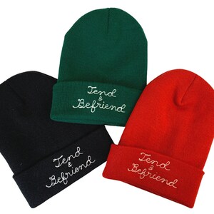 May include: Three knit beanies in black, green, and red. Each beanie has a folded cuff with the embroidered text "Tend & Befriend" in a cursive font. The beanies are displayed against a white background.