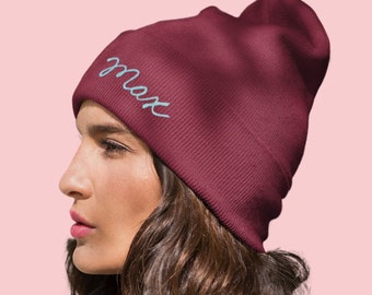 May include: A burgundy beanie hat with the name "Max" embroidered in light blue script. The hat is made of a ribbed knit material. The person wearing the hat has long, wavy brown hair.