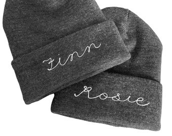 May include: Two dark gray knit beanies with white embroidered names. One beanie says "Finn" and the other says "Rosie" in a cursive font. The beanies are folded at the brim.
