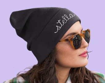 May include: A black knit beanie featuring the word stella embroidered in white cursive script on the folded cuff. The hat is made of a ribbed fabric and has a slouchy fit. The design is simple and features a clean, white thread embroidery.
