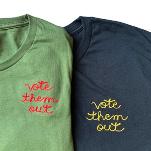 May include: Two t-shirts, one olive green and one navy blue, are displayed. Each shirt has the phrase "vote them out" embroidered on the front. The olive green shirt has red embroidery, while the navy blue shirt has yellow embroidery.