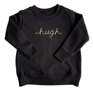 May include: Black crew neck sweatshirt with the name "hugh" embroidered in gold cursive script across the chest. The long-sleeved top has ribbed cuffs and a ribbed hem. The sweatshirt is made of a soft material.