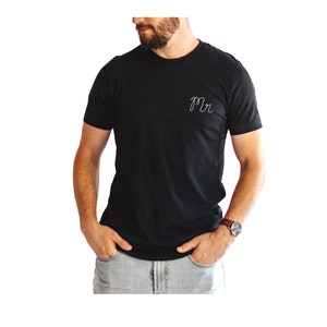 May include: Black crew neck t-shirt with the word "Mr" embroidered in white on the chest. The shirt is a solid color and has short sleeves. The model is wearing a watch with a brown leather band.