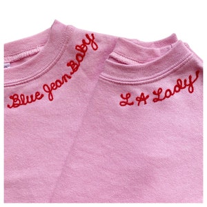 May include: Two light pink sweatshirts with red embroidered text. One says "Blue Jean Baby" and the other says "L.A. Lady". The sweatshirts are made of a soft material and have a crew neck design.