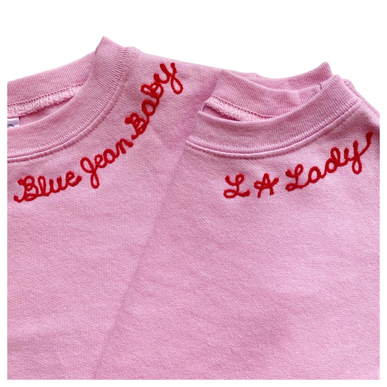 May include: Two light pink sweatshirts with red embroidered text. One says "Blue Jean Baby" and the other says "L.A. Lady". The sweatshirts are made of a soft material and have a crew neck design.