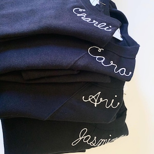 May include: A stack of navy blue sweatshirts embroidered with names in white cursive script. The names "Charlie", "Cara", "Ari", and "Jasmine" are visible. The sweatshirts are neatly folded, showcasing the personalized embroidery.