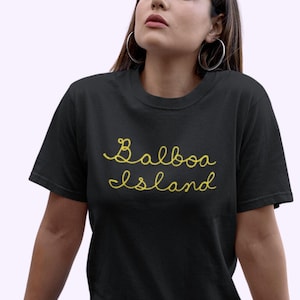 May include: Black t-shirt featuring the words "Balboa Island" in yellow script. The top has a crew neck and short sleeves. The text is centered on the chest.