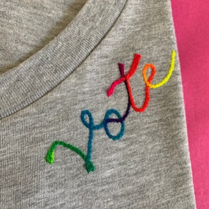 May include: A grey V-neck t-shirt with the word "vote" embroidered in rainbow colours. The embroidery is in a cursive style, with each letter in a different colour of the rainbow.