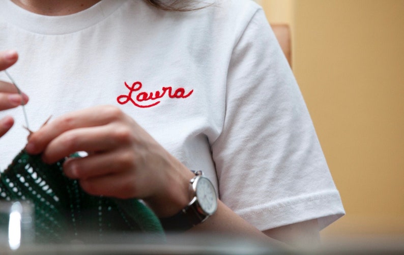 May include: White t-shirt with the name "Laura" embroidered in red script. The person is wearing a watch with a silver case and a black leather band. The person is knitting with green yarn.