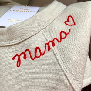 May include: A cream-coloured sweatshirt featuring the word "mama" embroidered in red script, with a small red heart. The garment has a crew neck and a tag that reads "CLUB CHAINSTITCH" and "handmade in Chicago."