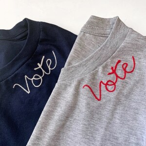 May include: Two t-shirts, one navy blue and one heather gray, are displayed. Each shirt has the word "Vote" embroidered on the chest in a cursive font. The navy shirt has white embroidery, while the gray shirt has red embroidery.