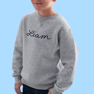 May include: A gray sweatshirt with the name "Liam" embroidered in black script across the chest. The sweatshirt has a crew neck and long sleeves. The background is a light blue.