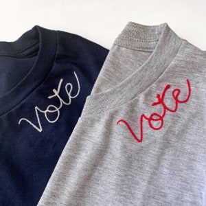 May include: Two t-shirts, one navy blue and one heather gray, are displayed. Each shirt has the word "Vote" embroidered on the upper chest. The navy shirt has white embroidery, while the gray shirt has red embroidery.