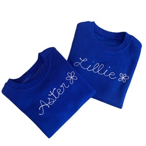 May include: Two royal blue sweatshirts with white embroidered names and flower accents. One sweatshirt says "Aster" and the other says "Lillie" in cursive script. The sweatshirts are displayed on a white background.