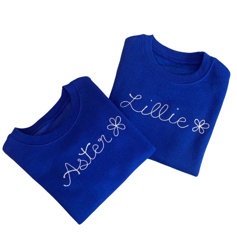 May include: Two royal blue sweatshirts with white embroidered names and flower accents. One sweatshirt says "Aster" and the other says "Lillie" in cursive script. The sweatshirts are displayed on a white background.