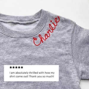 May include: A gray sweatshirt with the name "Charlie" embroidered in red script across the chest. The image includes a customer review with five stars and the text: "I am absolutely thrilled with how my shirt came out! Thank you so much!"