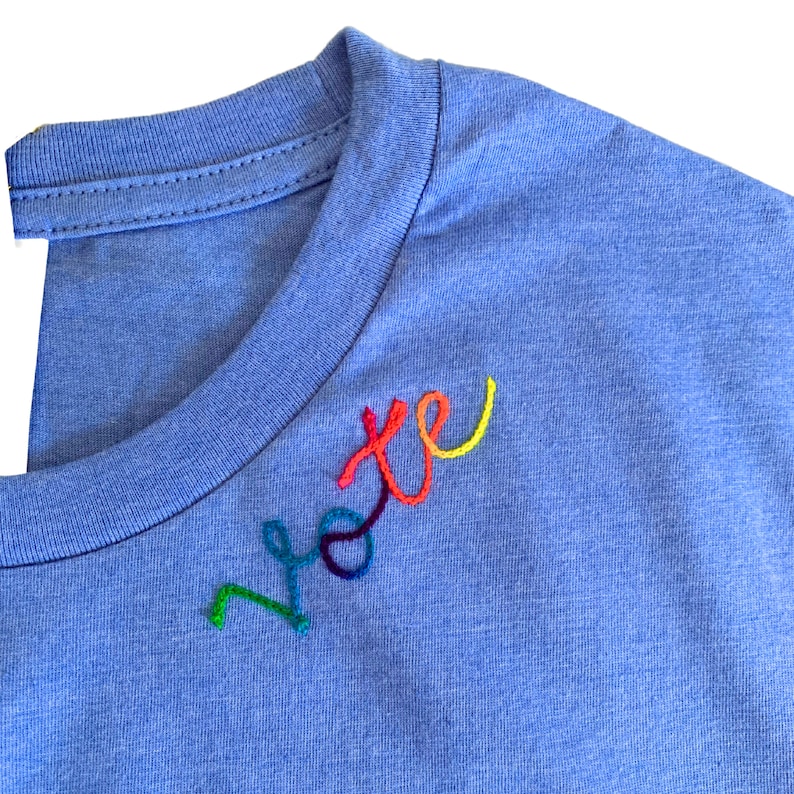 May include: A light blue t-shirt with the word "vote" embroidered in rainbow colors on the chest. The embroidery features a vibrant spectrum of hues, including green, yellow, red, and blue. The shirt has a crew neck.