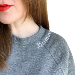 May include: A heather gray raglan sweatshirt with the word "Bummer" embroidered in white script on the shoulder. The sweatshirt has a crew neck and long sleeves. The model is wearing red lipstick.