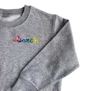 May include: A gray sweatshirt with the name "Sarah" embroidered in rainbow colors across the chest. The sweatshirt has a crew neck and long sleeves. The fabric appears to be a soft, knit material.