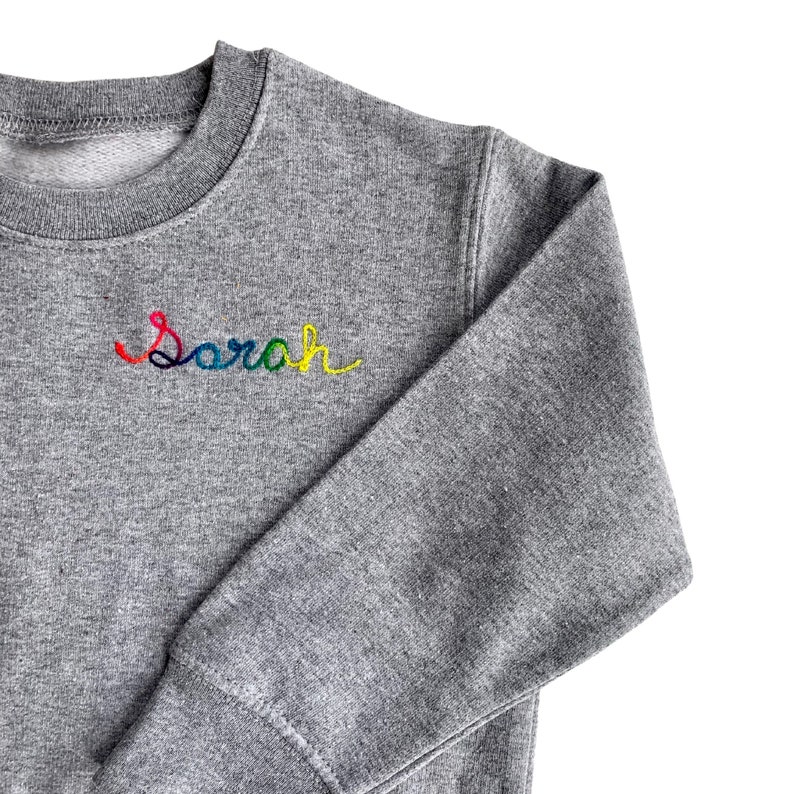 May include: A gray sweatshirt with the name "Sarah" embroidered in rainbow colors across the chest. The sweatshirt has a crew neck and long sleeves. The fabric appears to be a soft, knit material.