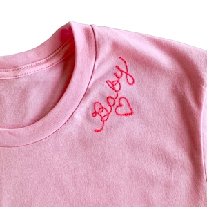 Puede incluir: Una camiseta de color rosa claro con texto bordado en rojo en letra cursiva que dice "baby" cerca del cuello, acompa&ntilde;ada de un peque&ntilde;o contorno de coraz&oacute;n rojo cosido a mano situado justo a la derecha de las letras bordadas.
