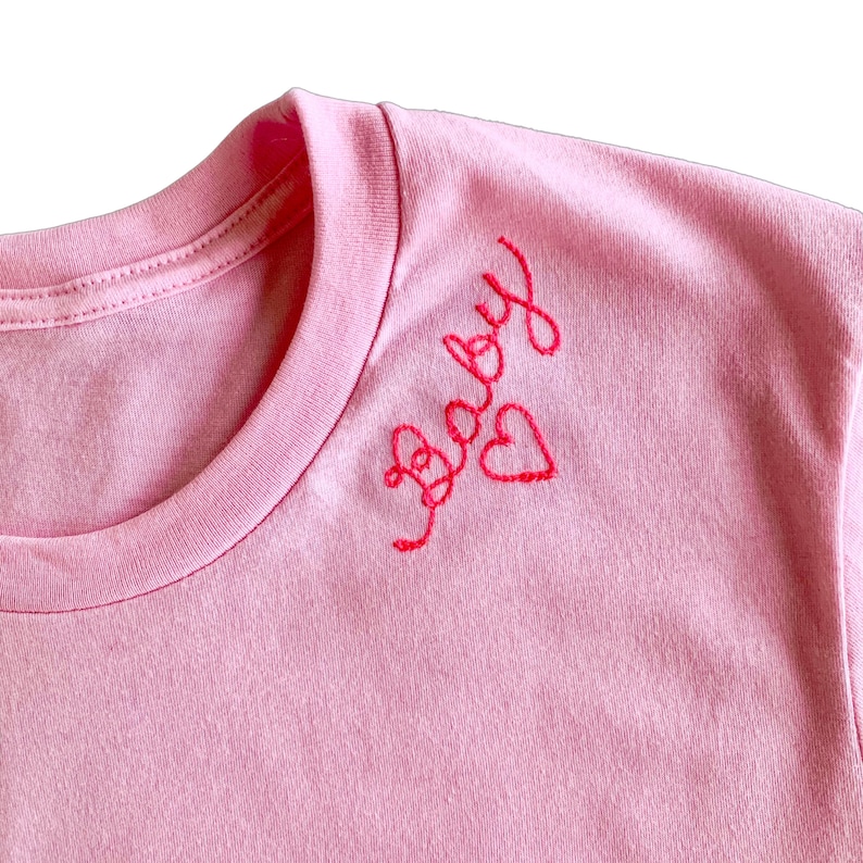 Puede incluir: Una camiseta de color rosa claro con texto bordado en rojo en letra cursiva que dice "baby" cerca del cuello, acompa&ntilde;ada de un peque&ntilde;o contorno de coraz&oacute;n rojo cosido a mano situado justo a la derecha de las letras bordadas.