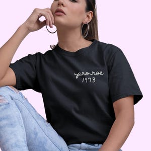 May include: Black t-shirt with the text "pro roe 1973" in white script. The shirt is a classic crew neck style. The model is wearing light blue ripped jeans.