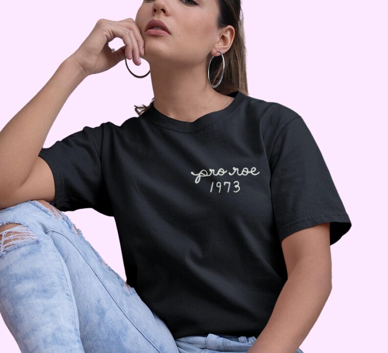 May include: Black t-shirt with the text "pro roe 1973" in white script. The shirt is a classic crew neck style. The model is wearing light blue ripped jeans.