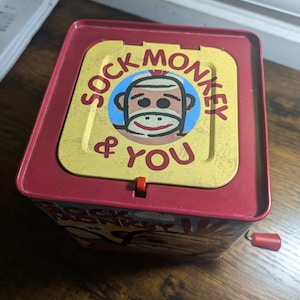 May include: A red and yellow metal Jack-in-the-Box toy with the words "SOCK MONKEY & YOU" printed on the lid. The lid features a cartoon sock monkey face. The toy has a red crank and a red latch.