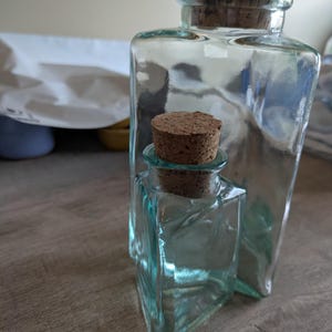 May include: Two clear glass bottles with cork stoppers. The bottles are different sizes and shapes, one square and one rectangular. The glass has a slight blue-green tint. The bottles are sitting on a wooden surface.