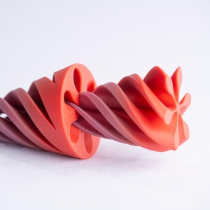 以下が含まれることがあります： 深い赤色から明るい赤紫色のグラデーションが美しい、3Dプリントされたオブジェクト。螺旋状のデザインで、湾曲した層状のセグメントが連なり、白い背景に配置されています。