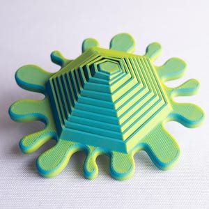 May include: A 3D-printed decorative object with a layered, geometric design. The central structure is a multi-tiered hexagon, surrounded by a splash-like border. The color scheme is a gradient of green and blue.