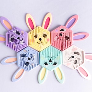 May include: A collection of colorful, hexagonal bunny-shaped decorations. Each bunny has a unique color and facial expression, with long ears extending outwards. The decorations are arranged in a circular pattern, creating a playful and festive design.