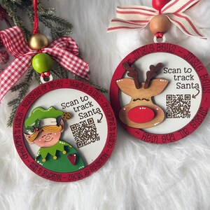 May include: Two round wooden Christmas ornaments with red borders. One features an elf, the other a reindeer. Both have a QR code with the text Scan to track Santa. The elf ornament says Naughty or Nice? Making a list, checking it twice. The reindeer ornament says Jingle Bells, Jingle all the way. Both have red and white ribbons.