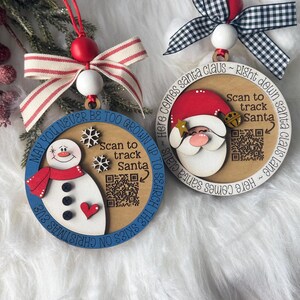 May include: Two round wooden ornaments with QR codes. One features a snowman with a red scarf and the text 'MAY YOU NEVER BE TOO GROWN UP TO SEARCH THE SKIES ON CHRISTMAS EVE'. The other shows Santa Claus with 'HERE COMES SANTA CLAUS ~ RIGHT DOWN SANTA CLAUS LANE'.