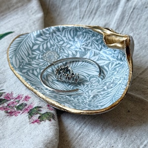 May include: A shell-shaped trinket dish with a gold rim and a blue and white floral pattern. A silver bracelet and a ring with dark stones are inside. The dish is displayed on patterned fabric. The dish is approximately 15 cm in length.