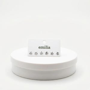 May include: A set of six small, silver-colored star-shaped stud earrings displayed on a white card with the brand name "emilia" printed in a black, sans-serif font. The earrings are on a white, round display.