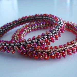 Op de afbeelding: Twee kralenarmbanden in roze en rode tinten met zilveren accenten. De armbanden zijn gemaakt van kleine, ronde kralen en zijn met elkaar verdraaid.