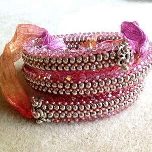 Puede incluir: Un brazalete de múltiples hilos hecho de cuentas plateadas y cristales rosas. El brazalete está envuelto con una cinta rosa y naranja.