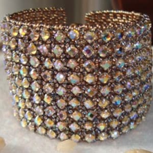 Puede incluir: Un brazalete ancho de color plateado con cuentas iridiscentes facetadas. El brazalete está hecho de muchas filas de cuentas, creando un aspecto texturizado y brillante.