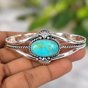 May include: A silver cuff bracelet featuring a central oval turquoise stone with brown matrix. The metal band has a twisted rope design surrounding the stone, flanked by two fan-shaped decorative elements at the top and bottom of the setting.